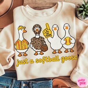 May include: Cream-colored sweatshirt with a playful design featuring four cartoon geese. The geese are dressed in softball-themed attire, including a jersey, a leopard-print uniform, and a foam finger. The text "just a softball goose" is printed below.