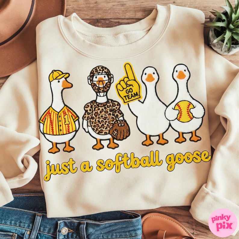 May include: Cream-colored sweatshirt with a playful design featuring four cartoon geese. The geese are dressed in softball-themed attire, including a jersey, a leopard-print uniform, and a foam finger. The text "just a softball goose" is printed below.