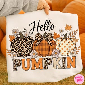 Op de afbeelding: Wit T-shirt met een herfstthema. Het shirt toont drie pompoenen met verschillende patronen, waaronder luipaardprint en stippen, samen met de woorden "Hello Pumpkin". Madeliefjes en herfstbladeren zijn ook inbegrepen.