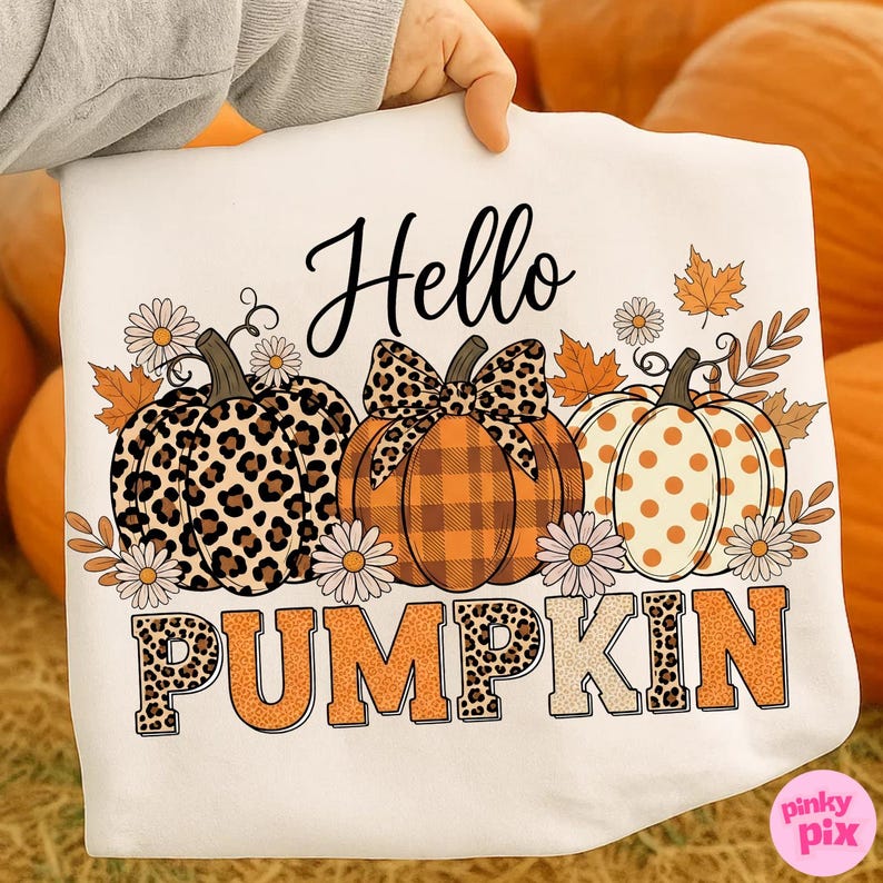 Op de afbeelding: Wit T-shirt met een herfstthema. Het shirt toont drie pompoenen met verschillende patronen, waaronder luipaardprint en stippen, samen met de woorden "Hello Pumpkin". Madeliefjes en herfstbladeren zijn ook inbegrepen.