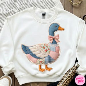 K&ouml;nnte beinhalten: Wei&szlig;es Sweatshirt mit einem gro&szlig;en, applizierten G&auml;nsemotiv. Die Gans hat einen blauen Kopf, einen orangefarbenen Schnabel und F&uuml;&szlig;e sowie eine rosa Schleife. Ihr K&ouml;rper ist ein Patchwork aus floralen, Vichy- und unifarbenen Stoffen, umrandet mit Perlen. Ein charmantes und skurriles Design.