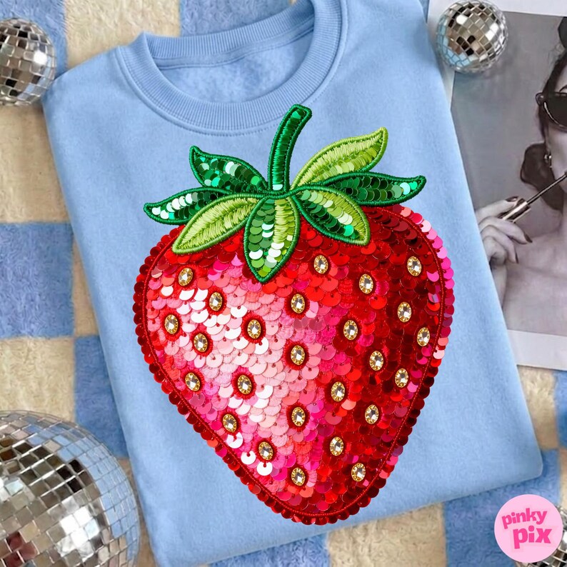 K&ouml;nnte beinhalten: Hellblaues Sweatshirt mit einem gro&szlig;en, paillettenbesetzten Erdbeermotiv. Die Erdbeere ist rot mit goldenen Akzenten und gr&uuml;nen Bl&auml;ttern. Das Sweatshirt hat einen Rundhalsausschnitt und lange &Auml;rmel. Das Pinky Pix-Logo ist unten rechts sichtbar.