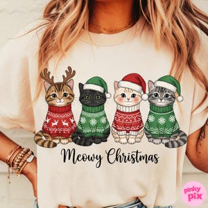 以下が含まれることがあります： クリスマスセーターと帽子をかぶった4匹の漫画の猫が描かれたクリーム色のTシャツ。猫は雪の結晶とトナカイのデザインの赤と緑のセーターを着ています。「Meowy Christmas」の文字が下に印刷されています。
