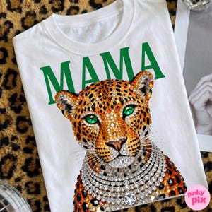 May include: White t-shirt with the word "MAMA" in green. It features a leopard graphic with green eyes and a diamond necklace. The leopard has a jeweled design.