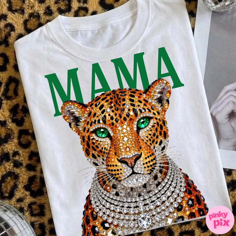May include: White t-shirt with the word "MAMA" in green. It features a leopard graphic with green eyes and a diamond necklace. The leopard has a jeweled design.