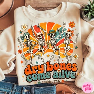May include: A cream-colored sweatshirt featuring a graphic of four dancing skeletons against a retro orange and teal sunburst. The skeletons wear accessories like a bandana, heart-shaped glasses, a hat, and a bow. Text reads dry bones come alive Ezekiel 37:5.