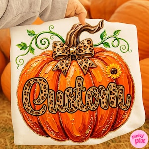 May include: A white t-shirt featuring a large orange pumpkin graphic with a brown stem, green vines, and a leopard print bow. The word Custom is written across the pumpkin in a cursive font with a leopard print pattern and gold glitter accents.