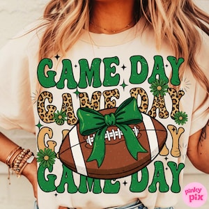 May include: Cream-colored t-shirt with a football graphic and the words "GAME DAY" in green, leopard print, and white. The football has a green bow. The shirt is a casual style.