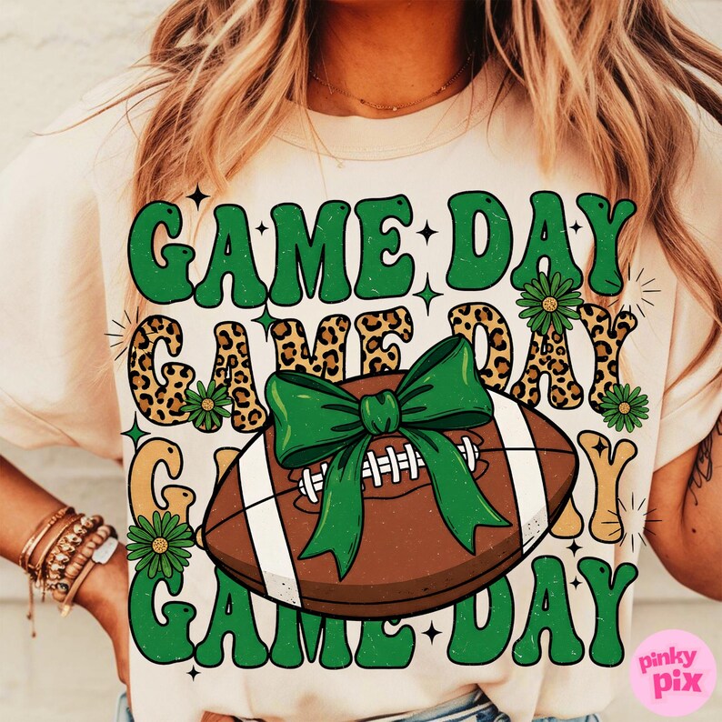 May include: Cream-colored t-shirt with a football graphic and the words "GAME DAY" in green, leopard print, and white. The football has a green bow. The shirt is a casual style.