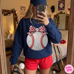 May include: Navy blue sweatshirt featuring a baseball design. The baseball is embellished with rhinestones and a red and white bow. The person is wearing red shorts and a navy blue baseball cap. A baseball glove and baseball are visible.