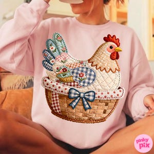 May include: A pale pink sweatshirt with a quilted chicken design in a basket. The chicken has a red comb and yellow beak. The basket has a blue bow. The chicken's feathers are a patchwork of floral and gingham patterns.