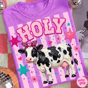 May include: A lilac t-shirt features a cow illustration with a leopard print bow and the word "HOLY" in pink bubble letters. The design includes pink stars, hearts, and flowers, creating a playful and whimsical aesthetic.