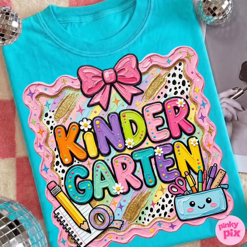 May include: A turquoise t-shirt with a colorful "Kindergarten" graphic. The graphic includes a pink bow, the word "Kindergarten" in various colors, and school supplies like a pencil, notebook, and scissors. The design is outlined in pink and gold.
