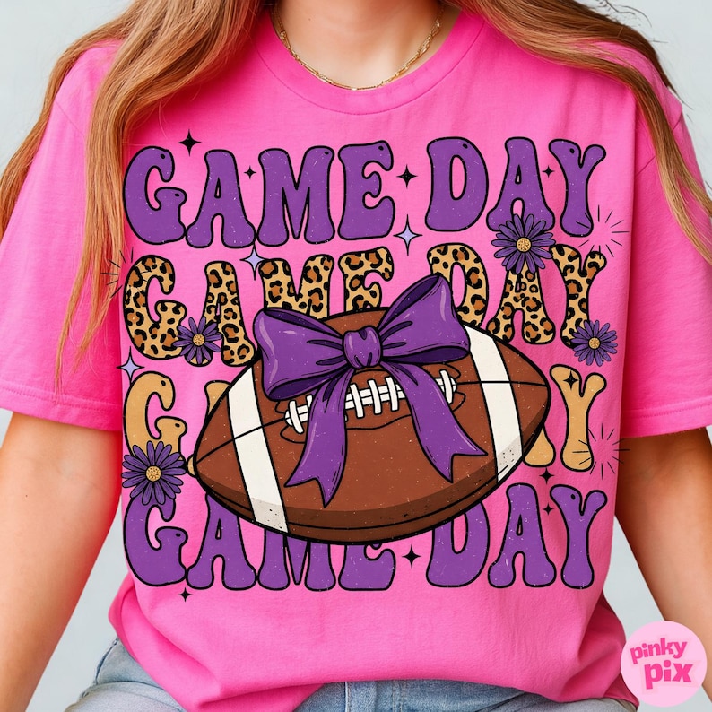 May include: A bright pink t-shirt with a retro design. The shirt features the words "GAME DAY" in purple, leopard print, and gold lettering. A football with a purple bow is in the center. Small purple flowers and stars complete the design.