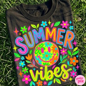 May include: Black t-shirt with a colorful "SUMMER VIBES" graphic. The design features a smiley face with flower details, surrounded by flowers and stars in bright neon colors. The shirt is displayed on a bed of green grass.
