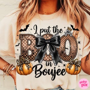 May include: Cream-colored t-shirt with a Halloween-themed graphic. The design features the phrase "I put the BOO in Boujee" with a large black bow, pumpkins, bats, and spiderwebs. The letters "B" and "O" have a checkered pattern.
