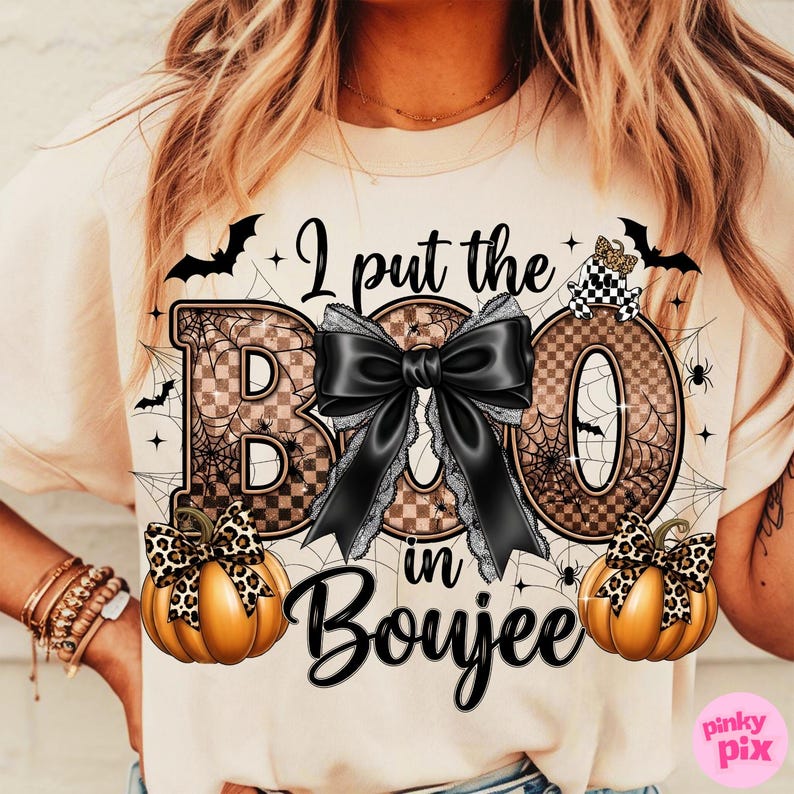 May include: Cream-colored t-shirt with a Halloween-themed graphic. The design features the phrase "I put the BOO in Boujee" with a large black bow, pumpkins, bats, and spiderwebs. The letters "B" and "O" have a checkered pattern.