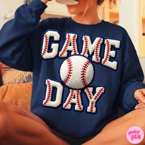 May include: Navy blue sweatshirt with the words "GAME DAY" in white, baseball-style lettering. A baseball graphic is centered on the sweatshirt. The lettering and baseball have red stitching details.
