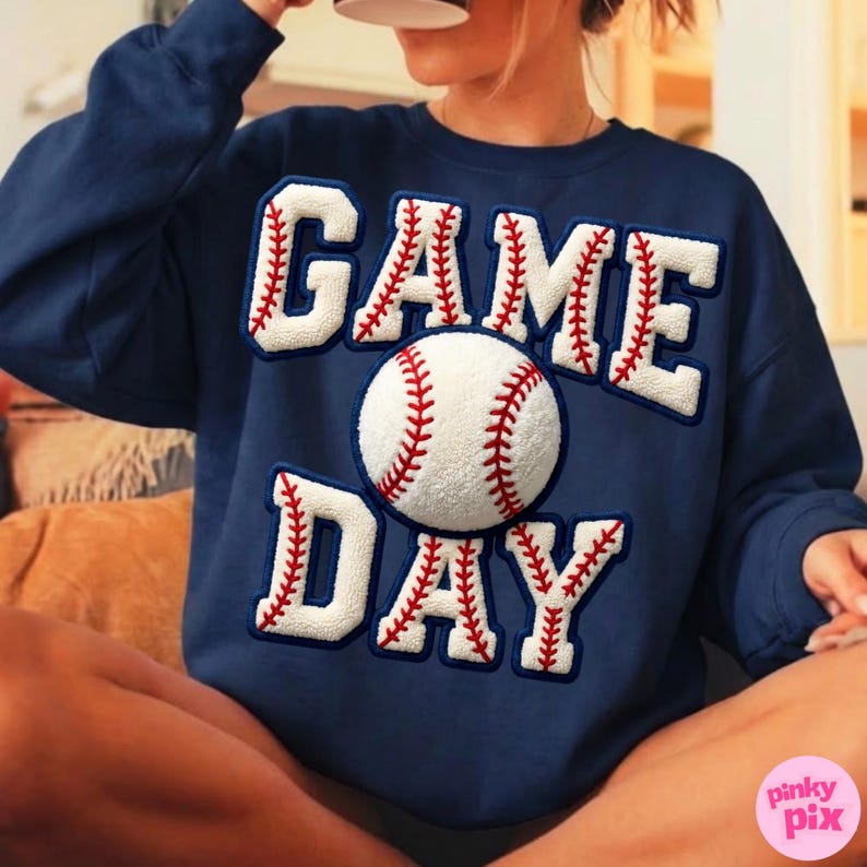 May include: Navy blue sweatshirt with the words "GAME DAY" in white, baseball-style lettering. A baseball graphic is centered on the sweatshirt. The lettering and baseball have red stitching details.