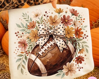 May include: A white sweatshirt featuring a graphic print of a brown football adorned with a large white bow with black polka dots, surrounded by a colorful arrangement of autumn-toned flowers in shades of orange, yellow, brown, and cream.