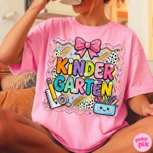 May include: Pink t-shirt with a colorful "Kindergarten" graphic. The graphic includes a pink bow, pencil, notebook, tape, scissors, and a pencil case. The shirt is a short-sleeved, oversized style.