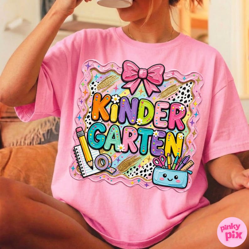 May include: Pink t-shirt with a colorful "Kindergarten" graphic. The graphic includes a pink bow, pencil, notebook, tape, scissors, and a pencil case. The shirt is a short-sleeved, oversized style.