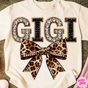May include: Cream-colored t-shirt featuring the word "GIGI" in large, jeweled letters. The letters are outlined in gold and filled with clear and black rhinestones. A large leopard print bow is centered below the text, embellished with gold accents.