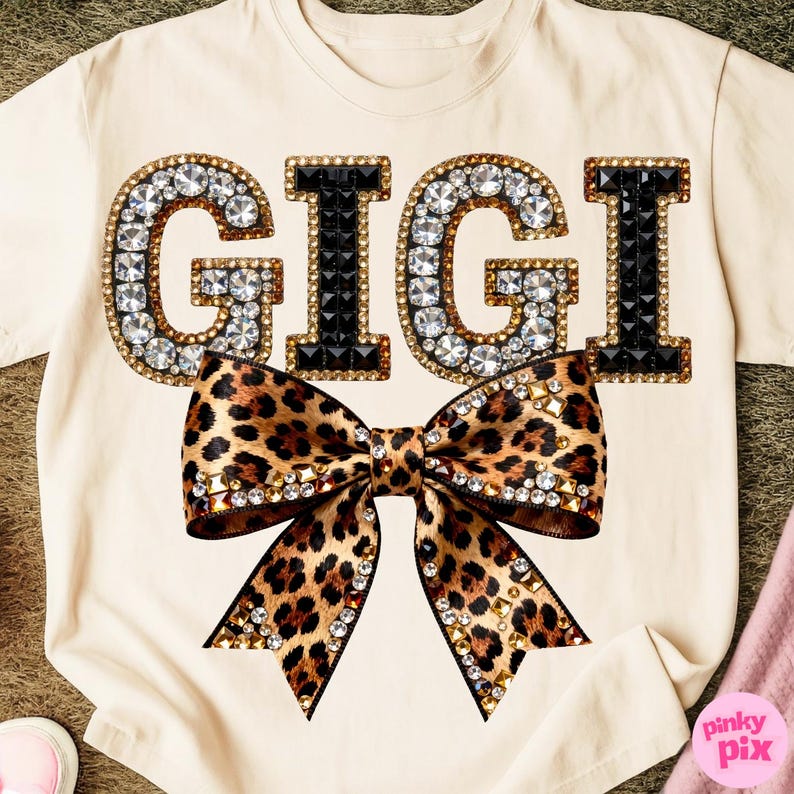 May include: Cream-colored t-shirt featuring the word "GIGI" in large, jeweled letters. The letters are outlined in gold and filled with clear and black rhinestones. A large leopard print bow is centered below the text, embellished with gold accents.
