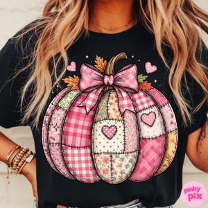 May include: Black t-shirt featuring a pink patchwork pumpkin design. The pumpkin has a pink bow, heart accents, and various patterned sections, including gingham and floral prints. The design is a fall or autumn theme.