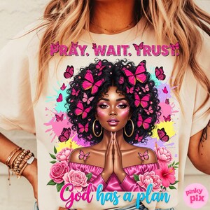 May include: A cream-coloured t-shirt with a colourful illustration of a woman adorned with butterflies. The text reads "Pray. Wait. Trust" and "God has a plan". The design incorporates pink, blue, and yellow splashes, creating a lively and artistic aesthetic.