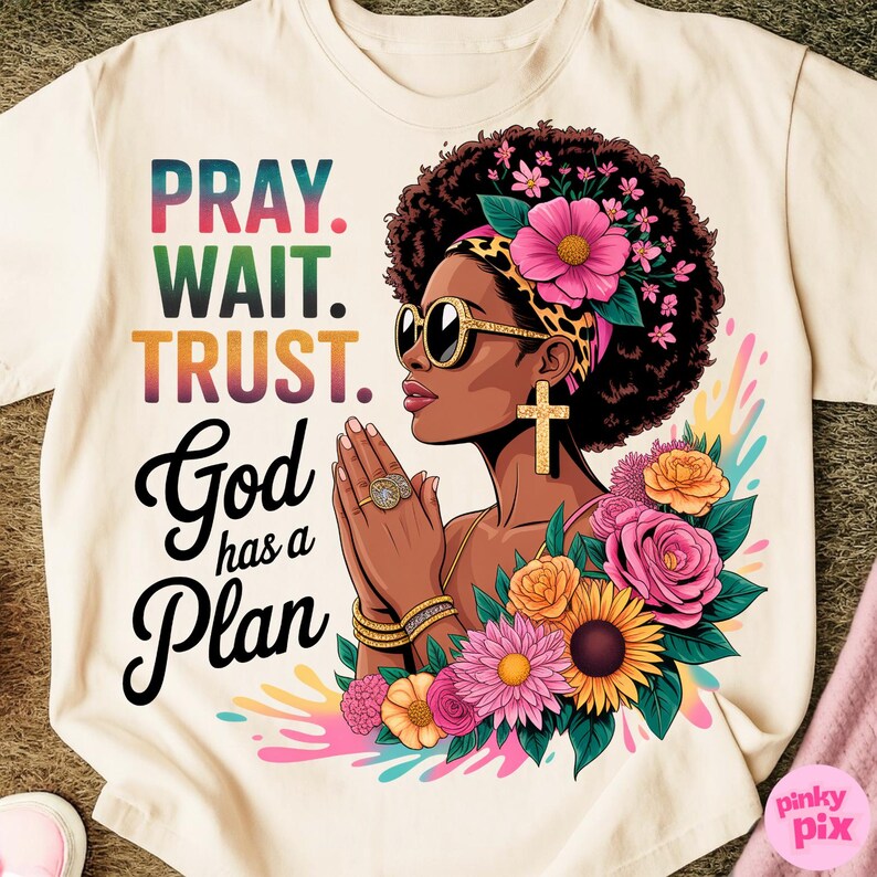 May include: Cream-colored t-shirt with the words "PRAY. WAIT. TRUST. God has a Plan" in colorful text. The graphic features a woman with sunglasses, a cross earring, and floral accents.
