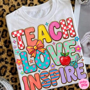 May include: White t-shirt with the words "TEACH LOVE INSPIRE" in colorful, bubble letters. The design includes a red apple, scissors, flowers, and a bow. The letters have a glittery gold outline.