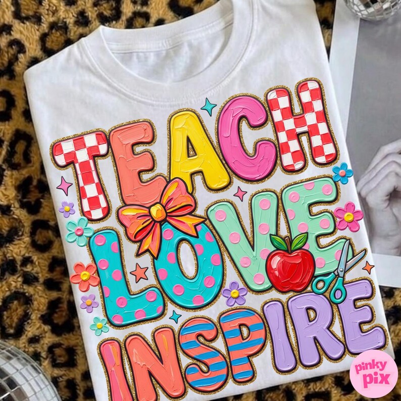 May include: White t-shirt with the words "TEACH LOVE INSPIRE" in colorful, bubble letters. The design includes a red apple, scissors, flowers, and a bow. The letters have a glittery gold outline.