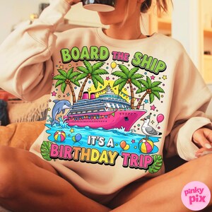 May include: A beige sweatshirt featuring a vibrant graphic of a pink cruise ship, palm trees, and a dolphin. The text reads "BOARD THE SHIP IT'S A BIRTHDAY TRIP". The design includes balloons, stars, and a seagull.