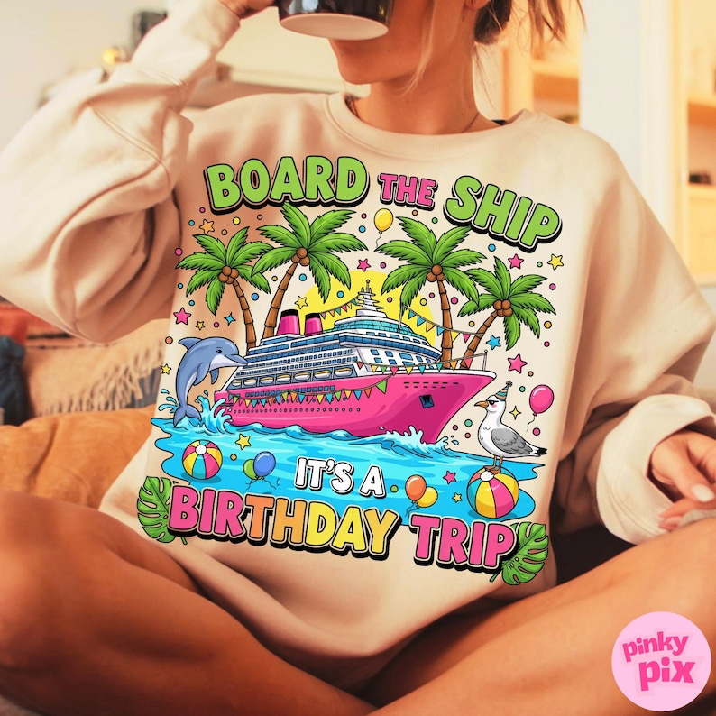 May include: A beige sweatshirt featuring a vibrant graphic of a pink cruise ship, palm trees, and a dolphin. The text reads "BOARD THE SHIP IT'S A BIRTHDAY TRIP". The design includes balloons, stars, and a seagull.