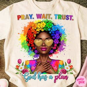 May include: Cream-colored t-shirt featuring a vibrant illustration of a woman with a floral afro and heart-shaped sunglasses. The shirt has the text "PRAY. WAIT. TRUST." at the top and "God has a plan" at the bottom, with a rainbow-colored background.