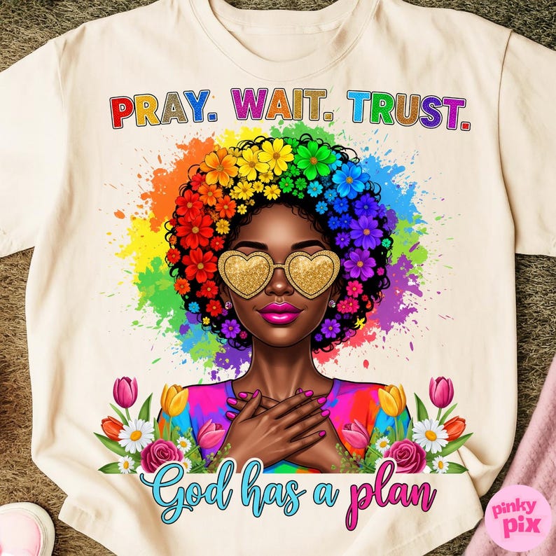 May include: Cream-colored t-shirt featuring a vibrant illustration of a woman with a floral afro and heart-shaped sunglasses. The shirt has the text "PRAY. WAIT. TRUST." at the top and "God has a plan" at the bottom, with a rainbow-colored background.