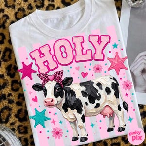 May include: White t-shirt featuring a cartoon cow with black and white spots, wearing a pink bow and the word "HOLY" in pink letters. The design includes pink stars, hearts, and flowers.