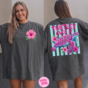 May include: Dark gray t-shirt with a pink hibiscus flower on the front. The back features the text "TURTLEY OVER IT" above a colorful turtle graphic with pink and blue accents, surrounded by flowers.