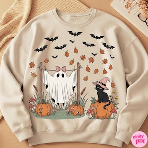 May include: Cream-colored Halloween-themed sweatshirt with a whimsical design. Features a ghost hanging on a clothesline, a black cat wearing a witch hat, pumpkins, bats, and falling leaves. The design is in shades of orange, black, and pink.