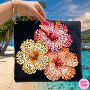 May include: Black square pillow featuring three sparkling hibiscus flowers in shades of red, orange, and yellow. The flowers are embellished with crystal-like accents. The background shows a tropical beach scene.