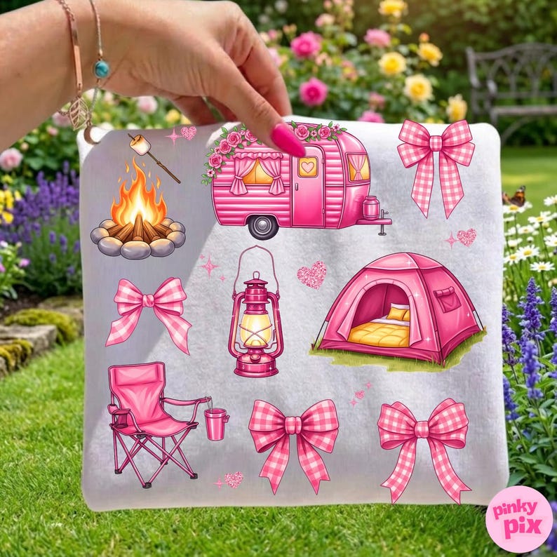 May include: A white camping-themed pillow featuring illustrations of a pink camper, tent, campfire, lantern, and bows. The design includes a pink folding chair and a marshmallow on a stick. The pillow is held in front of a green lawn and flower garden.