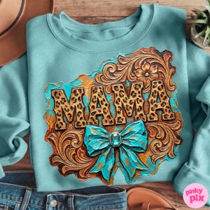 May include: A light blue sweatshirt with a decorative design. The design features the word "MAMA" in a leopard print pattern, set against a brown, ornate background with a turquoise bow.