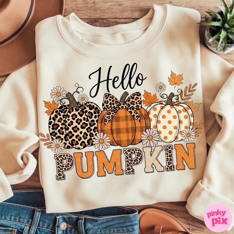 Op de afbeelding: Cr&egrave;mekleurige sweatshirt met een herfstthema. Het ontwerp toont drie pompoenen met verschillende patronen, waaronder luipaardprint en ruitjes, samen met de woorden "Hello Pumpkin". Het ontwerp bevat ook herfstbladeren en madeliefjes.