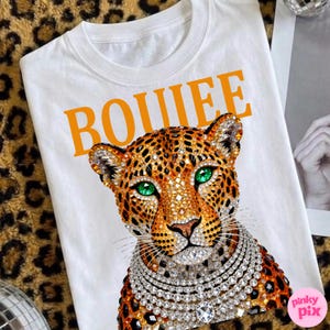 May include: White t-shirt with an orange "BOUIEE" text graphic. The shirt features a detailed leopard design with green eyes and a jeweled necklace. The leopard has a jeweled collar and is set against a leopard print background.