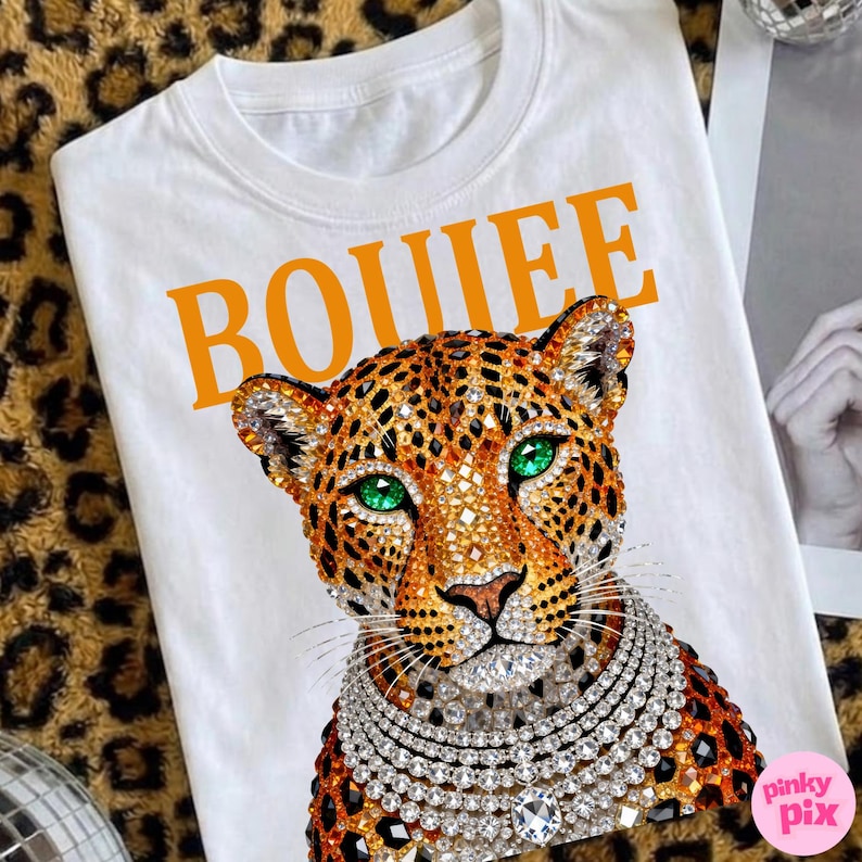 May include: White t-shirt with an orange "BOUIEE" text graphic. The shirt features a detailed leopard design with green eyes and a jeweled necklace. The leopard has a jeweled collar and is set against a leopard print background.