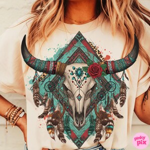 May include: A cream-coloured t-shirt with a detailed graphic of a bull skull decorated with turquoise and red accents, a red rose, and feathers. The design incorporates dream catchers and geometric patterns, evoking a bohemian aesthetic.