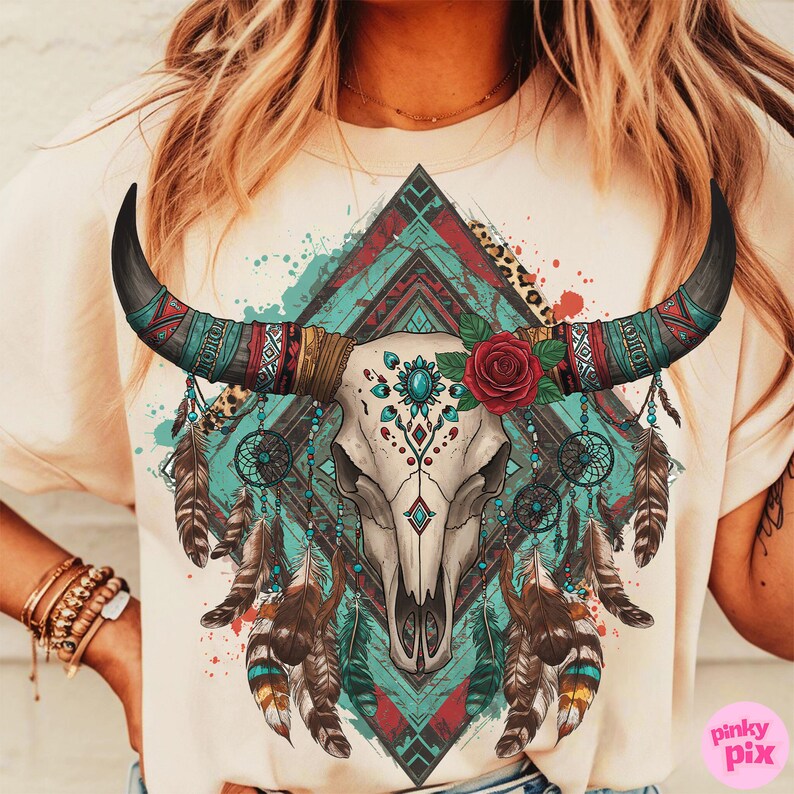 May include: A cream-coloured t-shirt with a detailed graphic of a bull skull decorated with turquoise and red accents, a red rose, and feathers. The design incorporates dream catchers and geometric patterns, evoking a bohemian aesthetic.