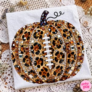 May include: A decorative pumpkin ornament featuring a leopard print pattern embellished with numerous brown, gold, and clear faceted crystals. A vertical white crystal-encrusted football lace design is centered on the front of the pumpkin.