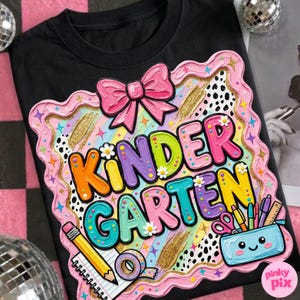 May include: Black t-shirt with a colorful "KINDERGARTEN" graphic. The design includes a pink bow, flowers, pencils, scissors, and a pencil case. The graphic has a textured, layered appearance.
